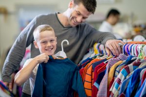 A little boy and his father are looking through a rack of clothes while shopping together. The boy has pulled out a long sleeve shirt to buy.